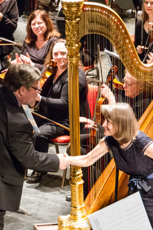 Ann with conductor Ann Lobotzke stands next to her harp and shakes a conductor's hand while members of the orchestra look on.